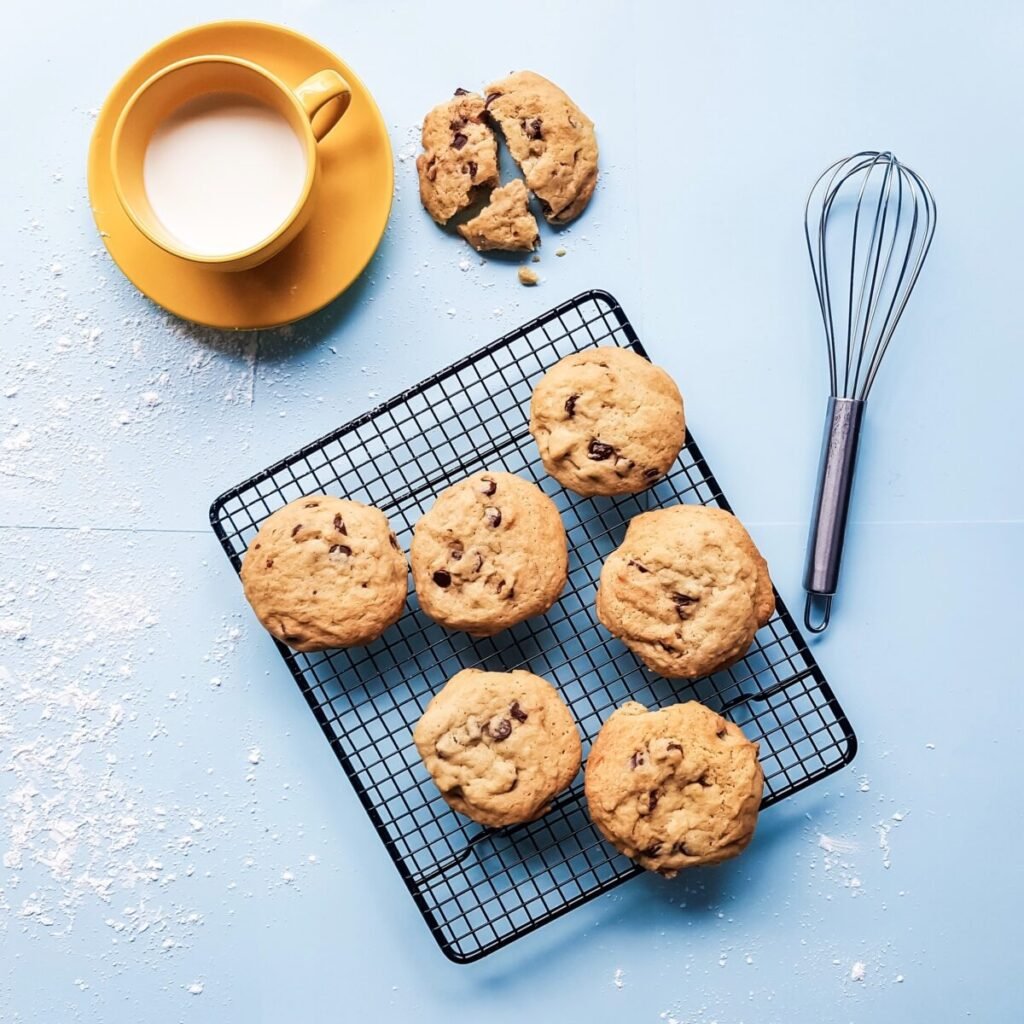 a cooling tray of fresh chocolate chip cookies, milk in a tea cup, and a wisk.