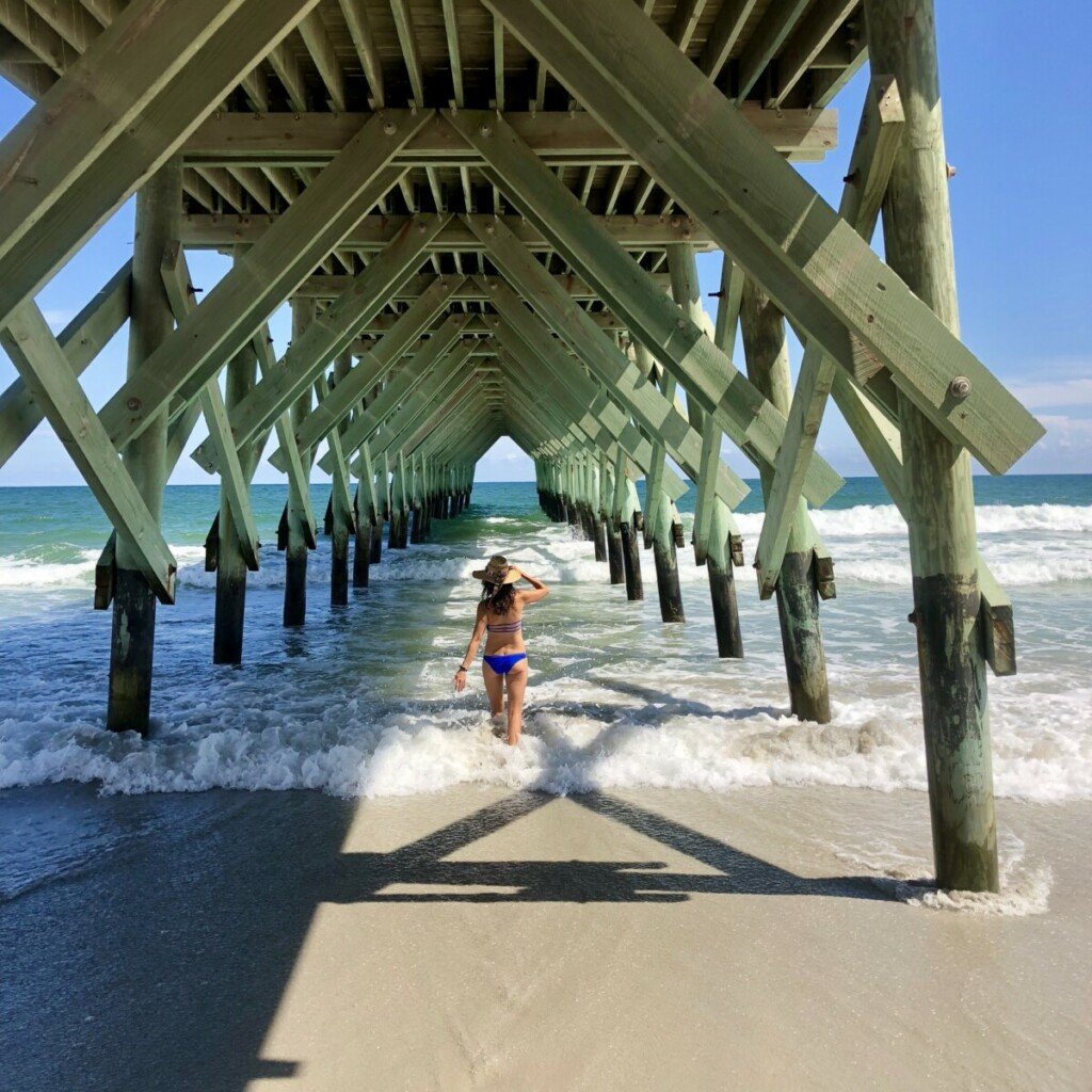 Woman walking under pier in turquoise water at Wrightsville Beach, North Carolina.