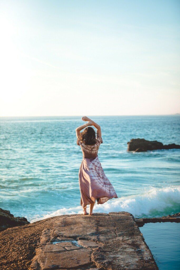 Woman in tye dye dress standing on a rock near the ocean with hands over her head.