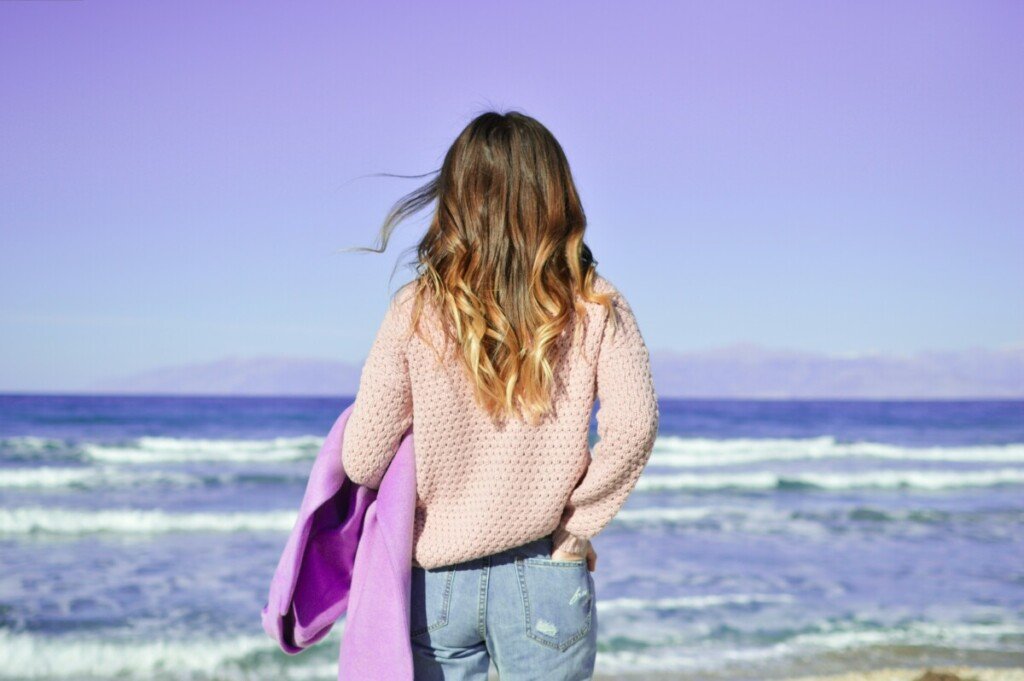 Woman with long brown hair and sweater and jeans looking toward the ocean
